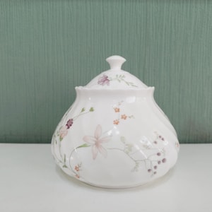 May include: White ceramic sugar bowl with a lid, decorated with delicate floral patterns in pink, purple, and orange. The bowl has a rounded shape and a small knob on the lid. The background is a solid teal colour.