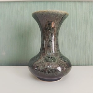 May include: A decorative vase with a flared top and a bulbous base. The vase has a dark, mottled glaze in shades of brown, green, and gray. The vase is sitting on a white surface against a light green wall.