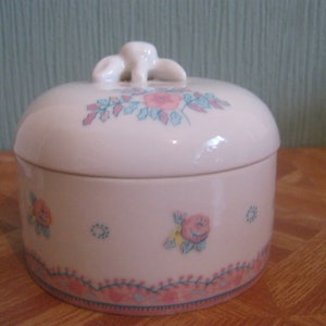 Könnte beinhalten: Eine weiße Keramikdose mit Blumenmuster und einem Deckel mit einem dekorativen Knopf. Die Dose hat ein rosa, blaues und grünes Blumenmuster.