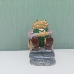 May include: A ceramic frog ornament seated on a stone-like base. The frog is tan with a green waistcoat, a lilac jacket, and brown shoes. The figurine has its hands clasped and appears to be smiling. The base is grey and textured.