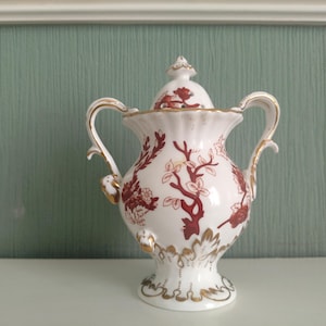 May include: A white porcelain teapot with a red and gold floral design. The teapot has a lid and two handles.
