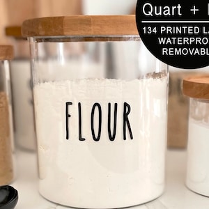 May include: Three glass jars with wooden lids and labels. The jars are filled with white powder. The labels read "Flour", "Sugar", and "Baking Soda".