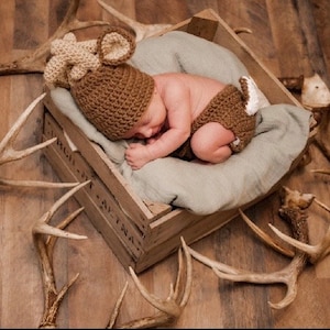 May include: A newborn baby wearing a brown and beige deer costume is sleeping in a wooden crate surrounded by antlers.
