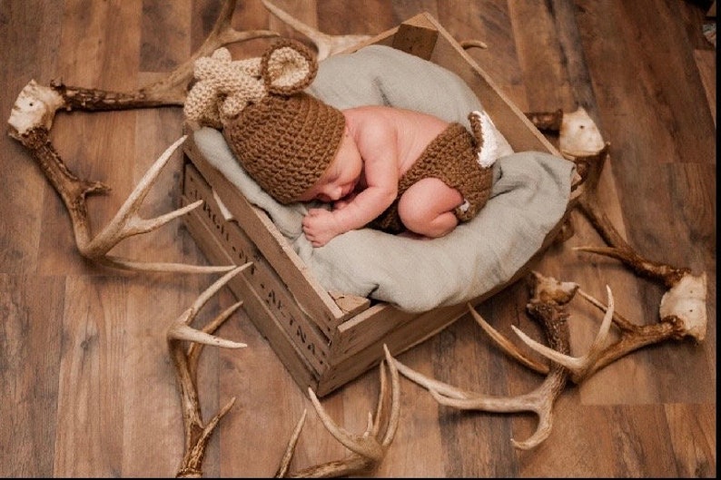 May include: A newborn baby wearing a brown and beige deer costume is sleeping in a wooden crate surrounded by antlers.