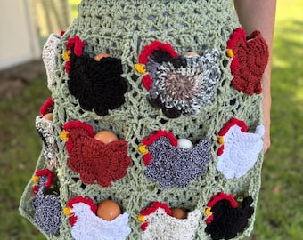 May include: A sage green crocheted apron featuring multiple pockets, each adorned with a small, colorful crocheted chicken applique. Several pockets contain brown or white chicken eggs. The apron has a grid-like pattern and a scalloped edge.