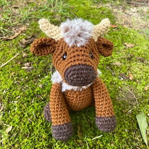 May include: A brown crocheted stuffed animal cow with a fluffy brown and white mane. The cow has brown eyes and a brown nose. The cow is sitting on a bed of green moss.