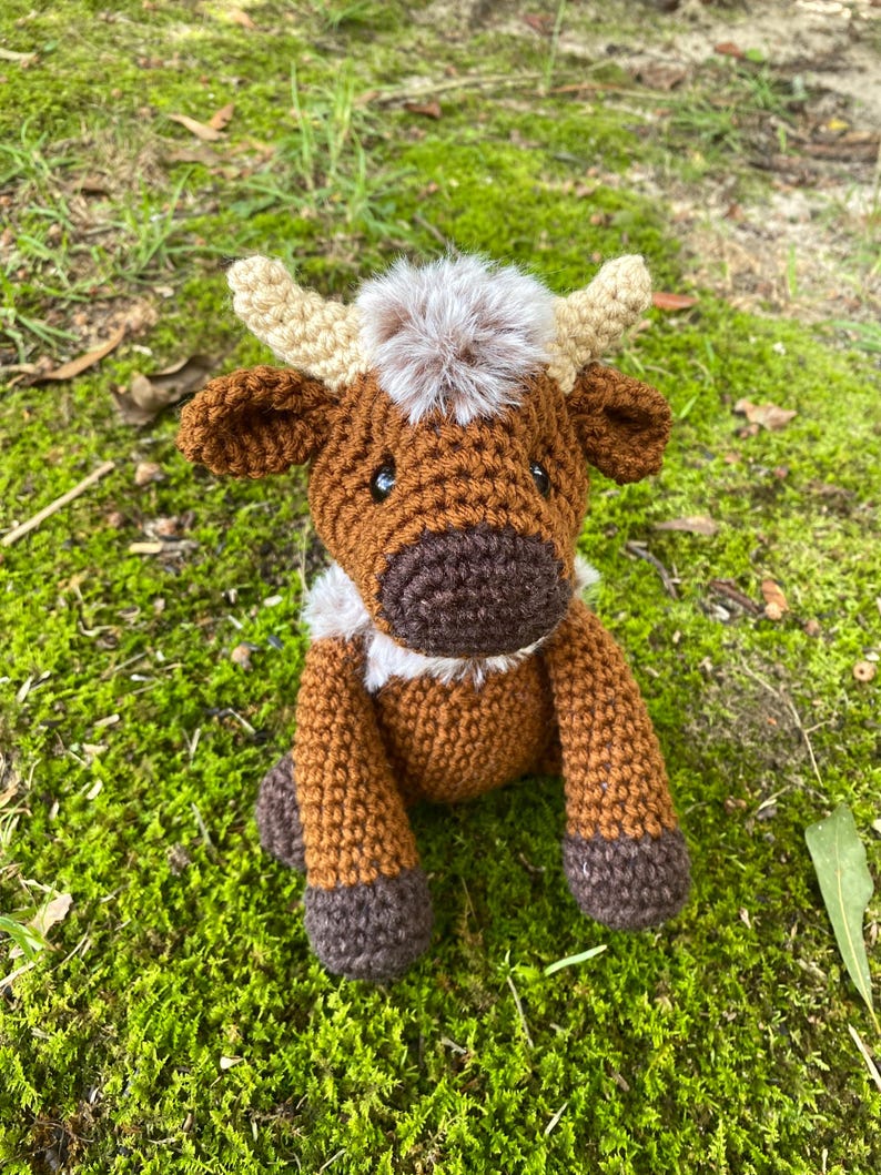 May include: A brown crocheted stuffed animal cow with a fluffy brown and white mane. The cow has brown eyes and a brown nose. The cow is sitting on a bed of green moss.