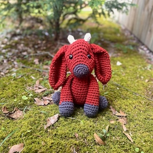 May include: A red crocheted goat toy with white horns and gray feet. The toy is sitting on a bed of green moss.