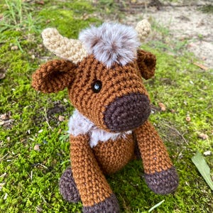 May include: A brown and white crocheted stuffed animal cow with a fluffy brown and white tail. The cow is sitting on a bed of green moss.