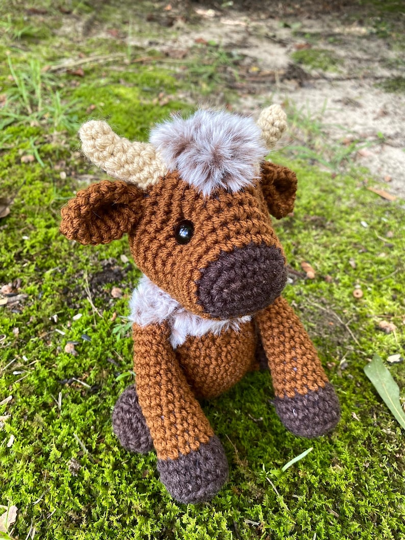 May include: A brown and white crocheted stuffed animal cow with a fluffy brown and white tail. The cow is sitting on a bed of green moss.