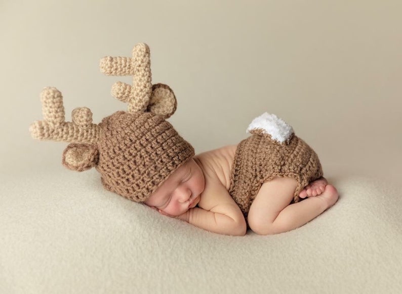 May include: A newborn baby wearing a brown crocheted reindeer hat and matching diaper cover with white accents. The baby is sleeping on a white fluffy surface.