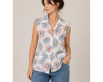 May include: White sleeveless button-down shirt with a colorful floral pattern. The shirt features a collared neckline and two front pockets. The floral design includes red, blue, yellow, and green accents. The shirt is paired with blue jeans.