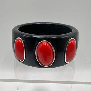 May include: A wide black bangle bracelet featuring three smooth, oval-shaped red cabochons set in silver-toned bezels. The bracelet has a polished, glossy finish and a uniform circular shape, showcasing a bold contrast between the black and red.