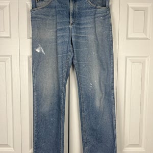 May include: Light blue denim jeans with a straight leg cut. The jeans have a classic five-pocket design, a button and zipper closure, and the Lee logo on the front pocket. There are white paint splatters on the front of the jeans.