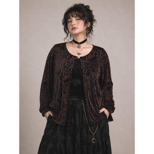 May include: A long-sleeved, open-front jacket in black velvet with a dark red paisley pattern. The jacket features a round neckline and is worn over a black top and a black tiered skirt. A silver chain with a crescent moon charm hangs from the waist.