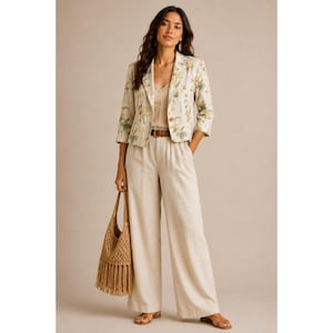 May include: A woman wearing a cream-colored floral print blazer with a matching top and wide-leg pants. She is also carrying a woven tote bag with fringe. The outfit is accessorized with a brown belt and sandals. The ensemble is suitable for spring or summer.