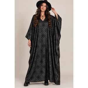 May include: A floor-length black kaftan dress featuring a tonal damask pattern, a V-neckline, and wide, flowing sleeves. The garment is paired with a black wide-brimmed hat and black ankle boots, creating a cohesive, dark-toned ensemble.