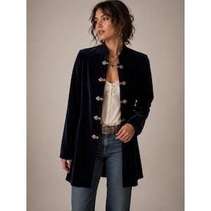 May include: A dark navy blue velvet jacket featuring a high collar and a front closure with five ornate, heart-shaped gold-tone metal clasps. The jacket has long sleeves and a straight hemline, worn over a white lace-trimmed top and blue denim jeans.