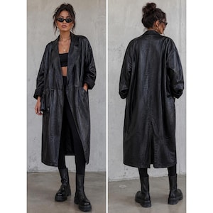 May include: A long black trench coat featuring a subtle snakeskin-like texture, a wide lapel collar, and long sleeves. The coat is worn open over a black top and black leggings, paired with black ankle boots, shown from both front and back views.