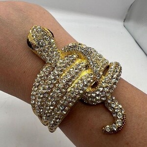 May include: A gold-toned snake bracelet featuring a coiled design covered in clear, sparkling rhinestones. The snake head is detailed with dark, prominent eyes, and the entire surface of the metal body is encrusted with small, reflective crystals.