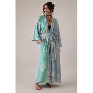 May include: A floor-length robe in shades of light blue and mint green, featuring a repeating sunburst pattern. The garment has wide, loose sleeves, a matching fabric tie belt at the waist, and a contrasting patterned border along the front opening.