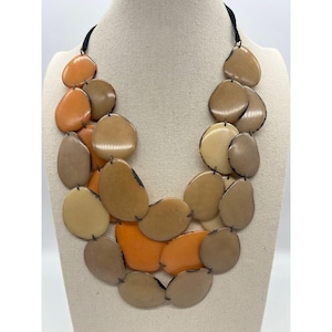 May include: A multi-layered statement necklace featuring smooth, rounded, pebble-like discs in shades of orange, tan, beige, and brown. The discs are connected by thin dark metal links and suspended from a simple black cord, creating a layered look.