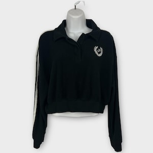 May include: Black cropped sweatshirt with a collared neckline and long sleeves. The sweatshirt features white stripes down the sleeves and a white embroidered emblem on the chest. The garment is displayed on a white mannequin.