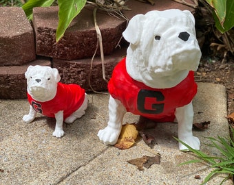 May include: Two white bulldog figurines wearing red and black jerseys with a large "G" on the front. The larger bulldog is sitting on a gray concrete surface with some fallen leaves around it. The smaller bulldog is sitting on the left side of the image.
