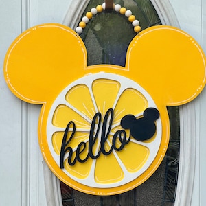 May include: A yellow lemon-shaped Mickey Mouse head wreath with the word "hello" in black lettering. The wreath is decorated with white and yellow beads.