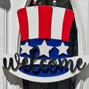 Könnte beinhalten: Ein rot-weiß-blaues patriotisches Willkommensschild in Form eines Zylinders mit Sternen und dem Wort "welcome" in schwarzen Buchstaben.