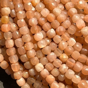 May include: A strand of peach-colored sunstone beads, each bead is faceted and has a square shape.