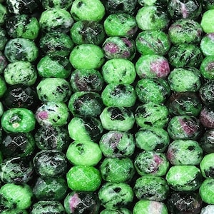 May include: A close-up of a strand of green, faceted gemstone beads. The beads have a speckled appearance with black and pink flecks throughout.