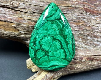 May include: A polished, teardrop-shaped malachite gemstone. The vibrant green stone features intricate swirling patterns and a small drilled hole at the top. The gemstone is displayed on a piece of weathered wood.