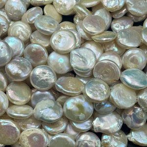 May include: A close-up of a pile of white, round, flat, iridescent pearls. The pearls are smooth and have a slightly irregular shape.