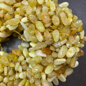 May include: A strand of polished, oval-shaped gemstones in shades of yellow, cream, and amber. The beads are strung together with gold-colored string, creating a necklace or bracelet. The gemstones vary in size and have a smooth, glossy finish.