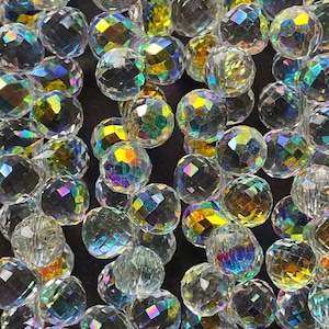 May include: Close-up of a collection of clear, faceted crystal beads. The beads have an iridescent finish, reflecting a spectrum of colors including yellow, green, and purple. The spherical beads are scattered, creating a textured, sparkling effect.