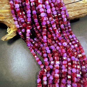 May include: Strands of faceted beads in shades of deep red and purple. The beads are cube-shaped and strung together, creating a textured and colorful display. The beads are displayed against a dark background.