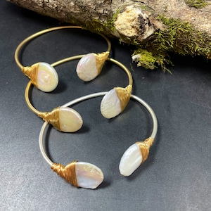 Pu&ograve; includere: Tre braccialetti avvolti in filo dorato e argentato con accenti di perle bianche. I braccialetti sono impilati su una superficie scura.
