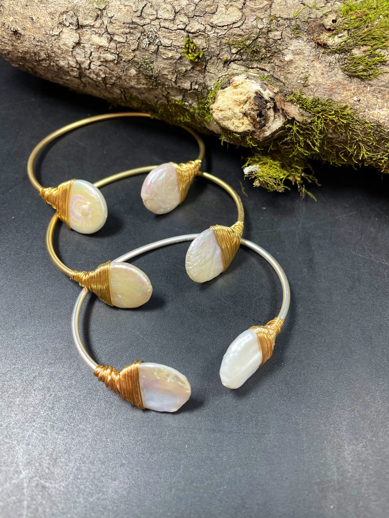 Pu&ograve; includere: Tre braccialetti avvolti in filo dorato e argentato con accenti di perle bianche. I braccialetti sono impilati su una superficie scura.