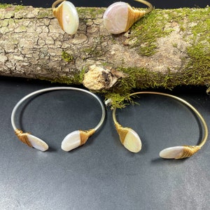 Pu&ograve; includere: Tre braccialetti avvolti in filo dorato con accenti di conchiglia bianca iridescente. I braccialetti sono esposti su una superficie di legno con muschio.