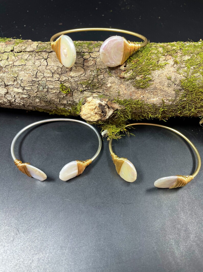 Pu&ograve; includere: Tre braccialetti avvolti in filo dorato con accenti di conchiglia bianca iridescente. I braccialetti sono esposti su una superficie di legno con muschio.