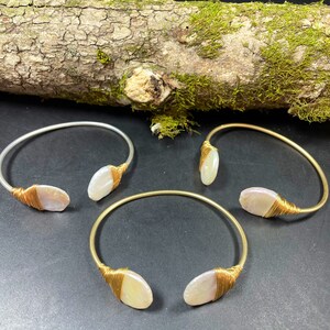 Pu&ograve; includere: Tre braccialetti avvolti in filo dorato con accenti di conchiglia bianca. I braccialetti sono su una superficie di legno con muschio.