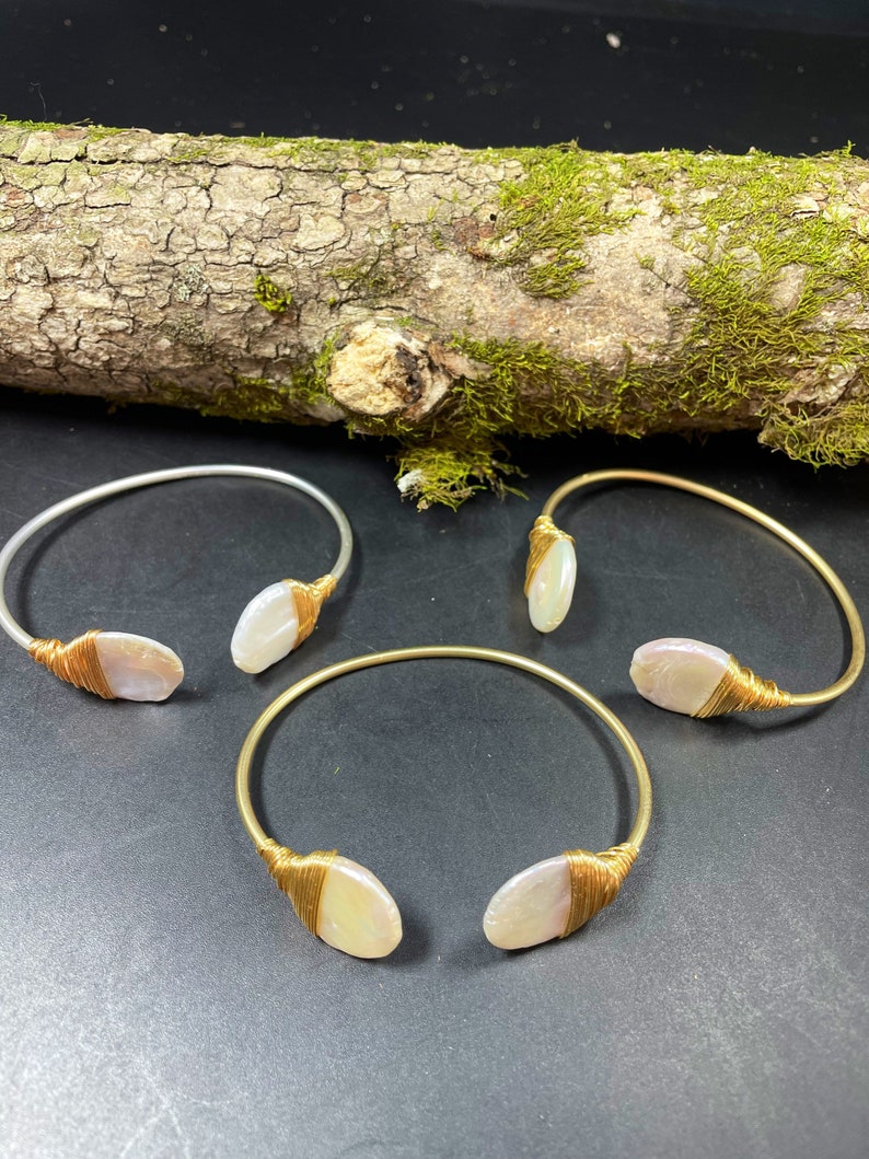 Pu&ograve; includere: Tre braccialetti avvolti in filo dorato con accenti di conchiglia bianca. I braccialetti sono su una superficie di legno con muschio.