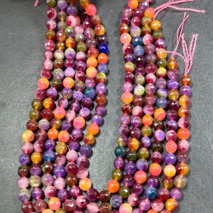 May include: Multicolored gemstone beads in shades of pink, purple, orange, and green. The beads are round and have a faceted finish. The beads are strung on a string and are ready to be used for jewelry making.