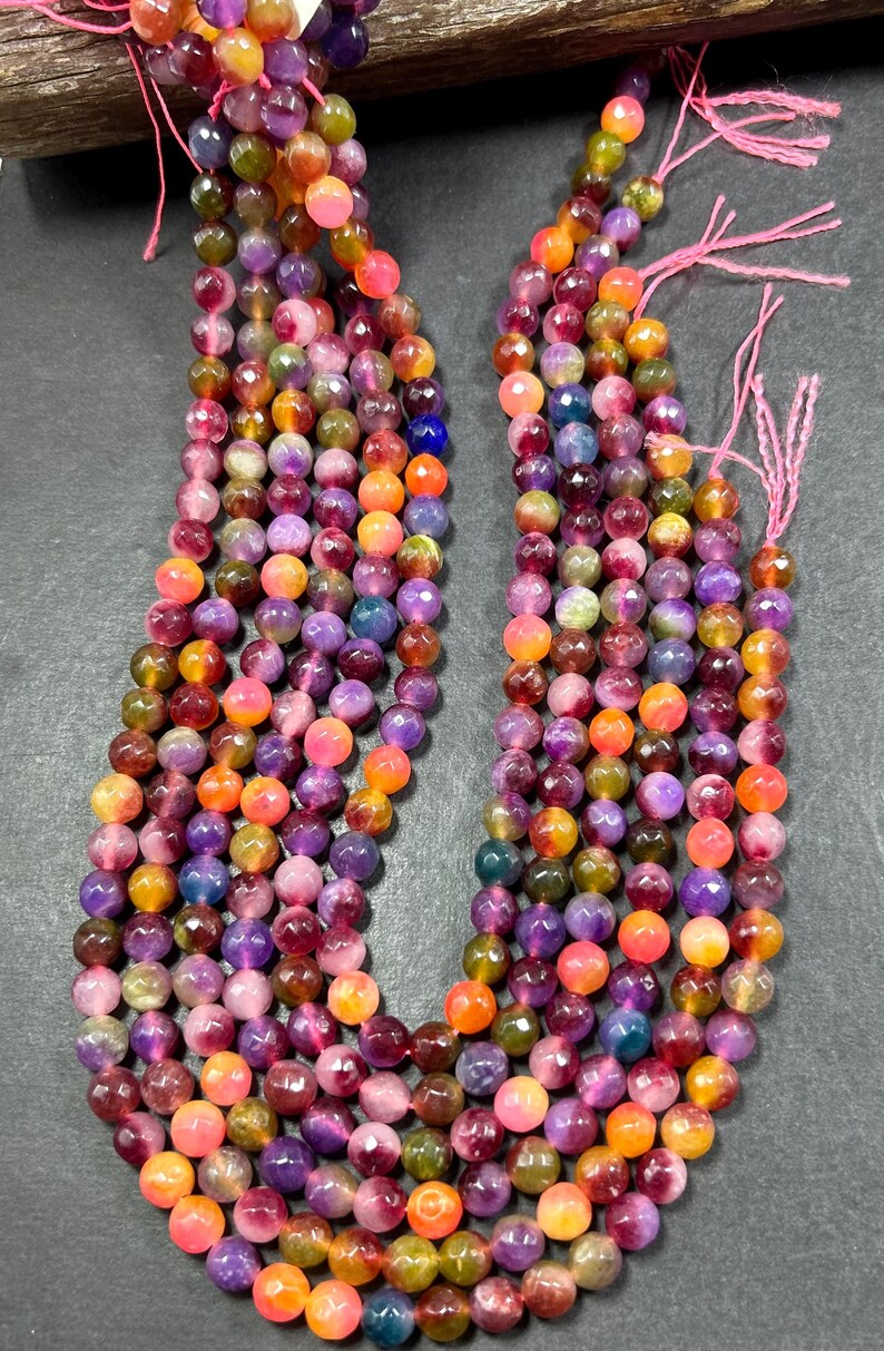 May include: Multicolored gemstone beads in shades of pink, purple, orange, and green. The beads are round and have a faceted finish. The beads are strung on a string and are ready to be used for jewelry making.