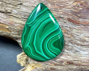 May include: A polished, teardrop-shaped malachite pendant with a drilled hole at the top. The gemstone features vibrant green hues with swirling patterns of darker green. It is set against a backdrop of weathered wood.