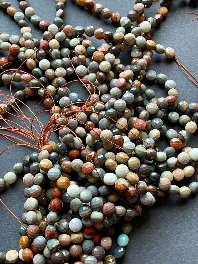 May include: A pile of multi-coloured gemstone beads with a faceted finish. The beads are strung on brown thread and are a mix of brown, red, green, and white.