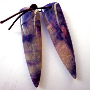 May include: Two elongated, pointed pendants made of polished stone with a smooth, glossy finish. The stones feature a marbled pattern of deep purple, translucent white, and soft pink hues. A thin, dark brown cord passes through a hole at the top.