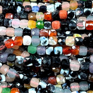 May include: Close-up of a collection of faceted gemstone beads in various colors. The beads are square-shaped and feature a range of colors including black, white, pink, green, orange, and blue. The beads are strung together, suggesting they are for jewelry making.