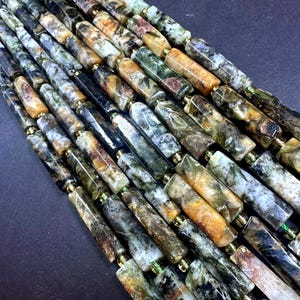 May include: A collection of cylindrical beads arranged in parallel strands. The beads feature a marbled pattern with variegated shades of forest green, earthy brown, cream, and black, with a polished finish and small gold-toned spacers.